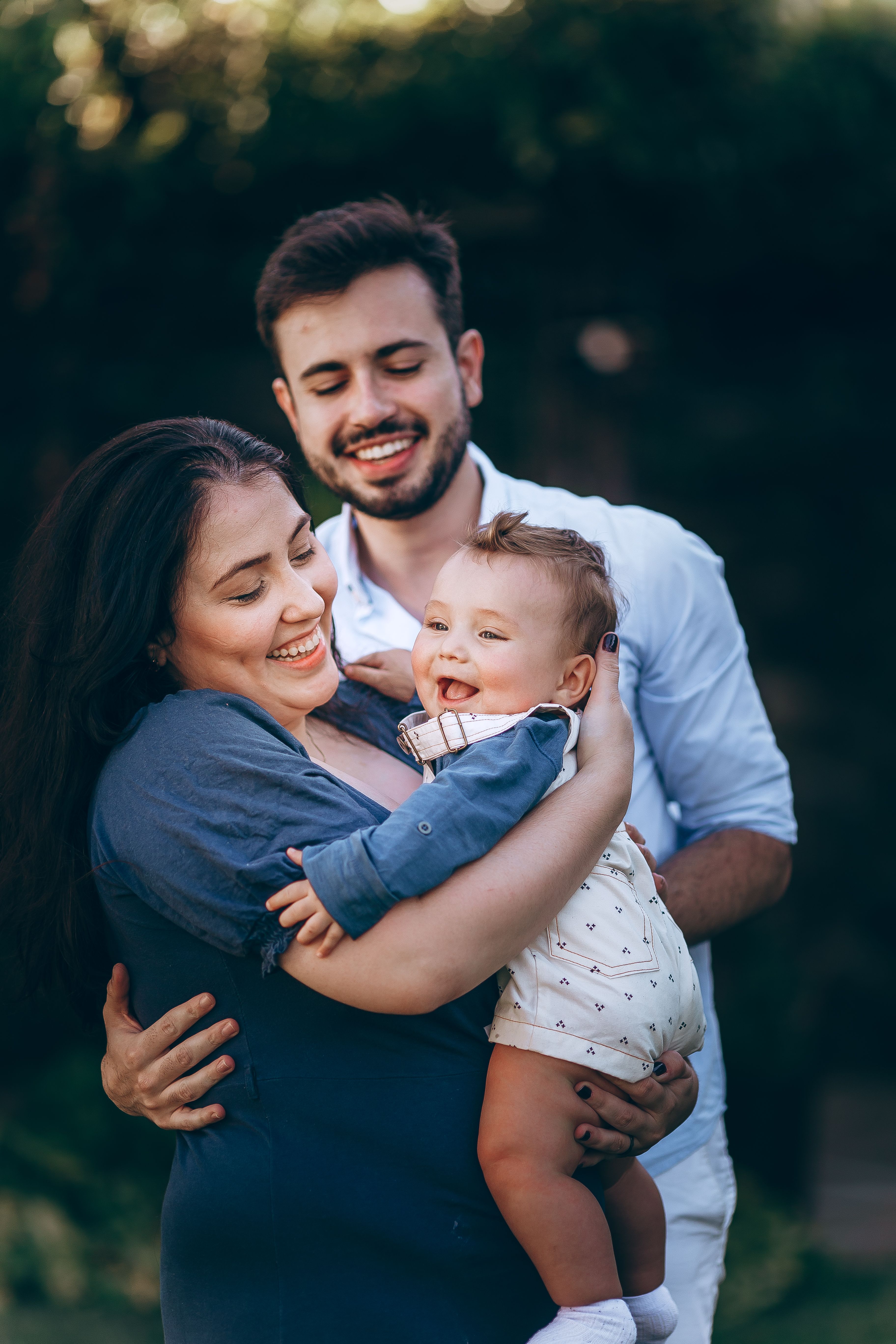 Two parents smiling outdoors while holding their baby.
