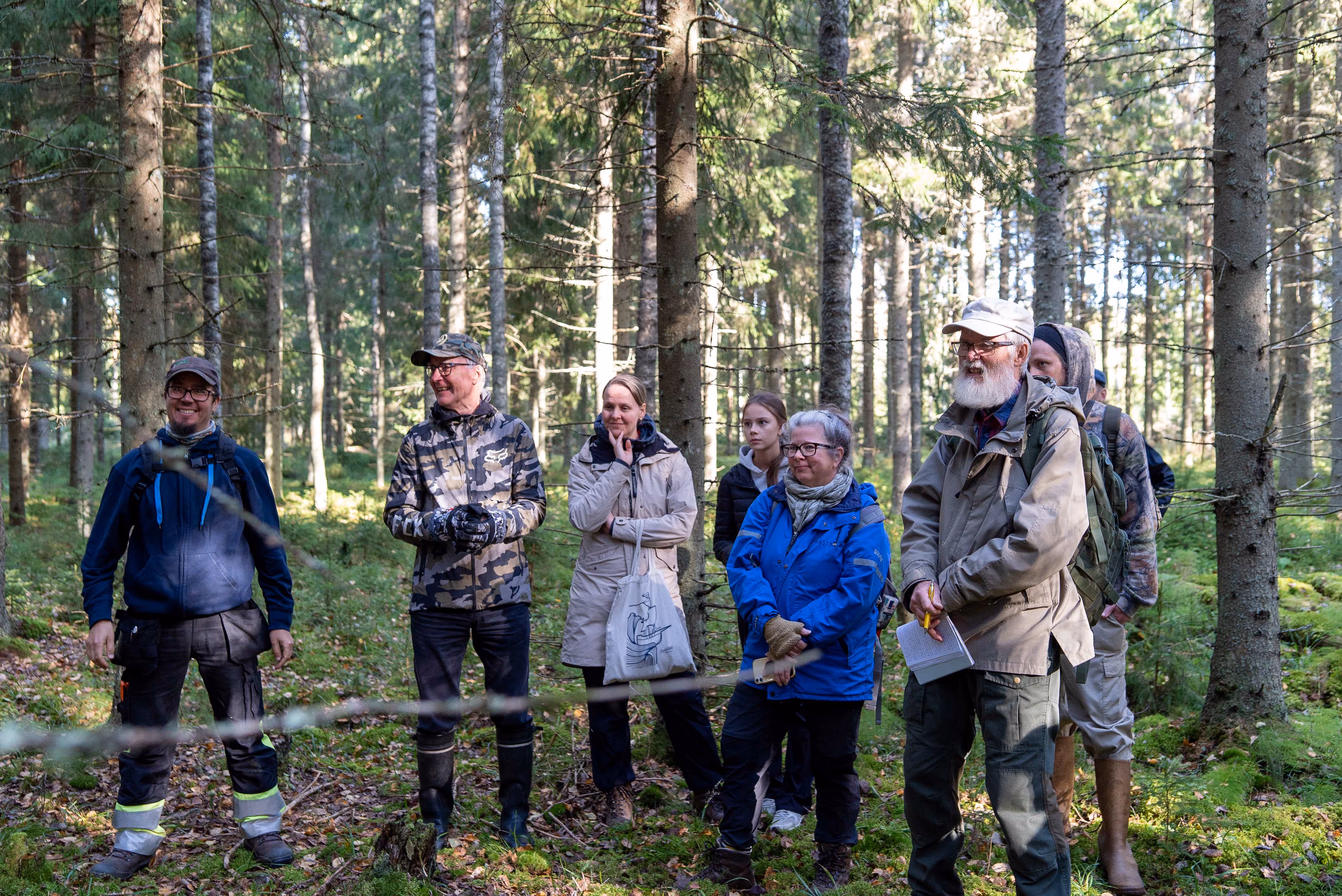 Ryhmän aikuisia ihmisiä metsässä aurinkoisena päivänä, osa seisoo hymyillen, osa kuuntelee.