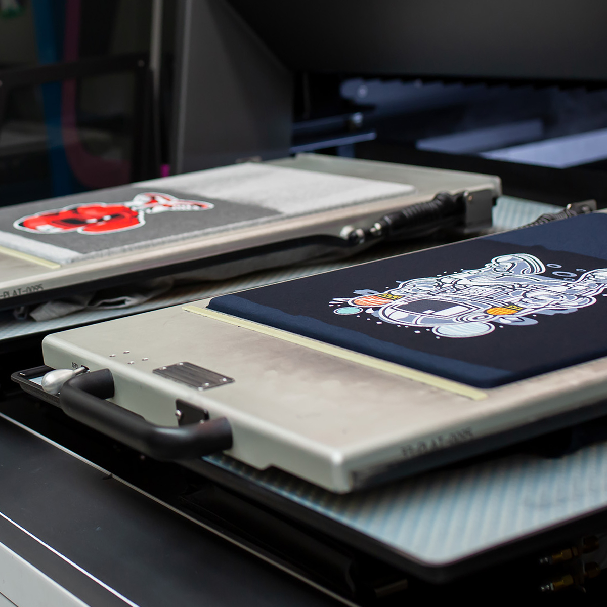 Close-up of a heat press machine with two fabric pieces, one black with a white graphic and one gray with a red graphic.