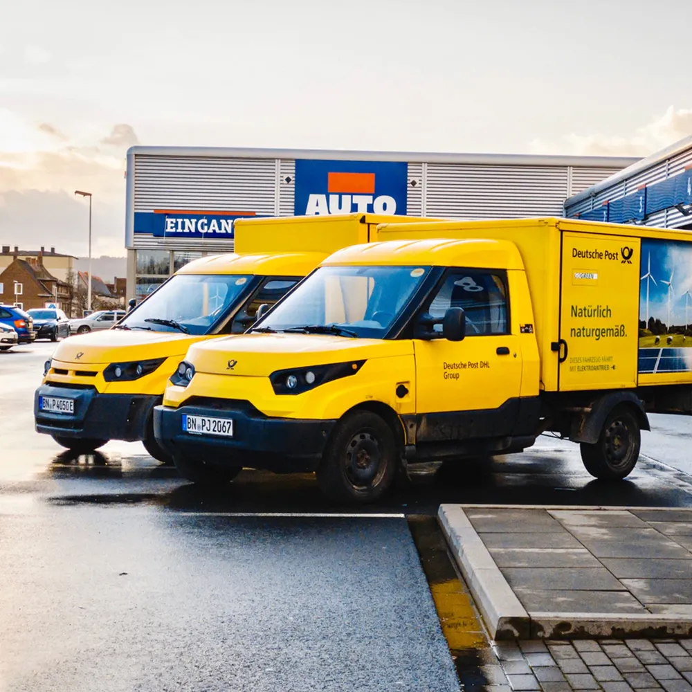 Two yellow Deutsche Post DHL delivery trucks parked outside an auto center with signage in German.
