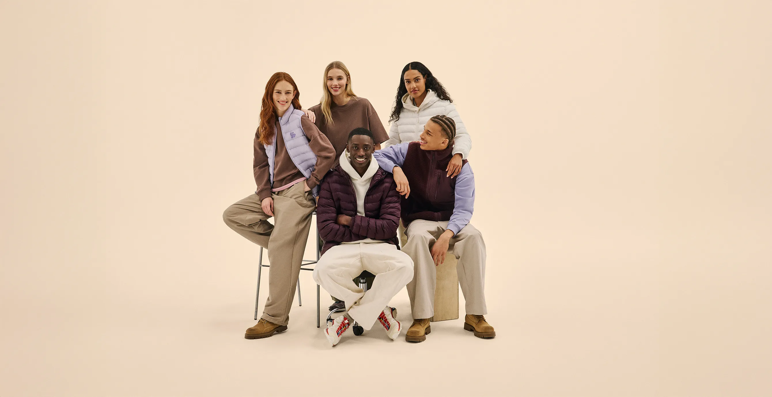 Five young adults smiling and posing in casual winter clothing against a beige background, with two seated and three standing behind them.
