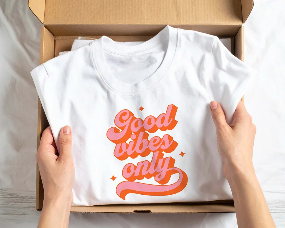 Hands holding a folded white t-shirt with orange and pink lettering that says 'Good vibes only' inside an open cardboard box.