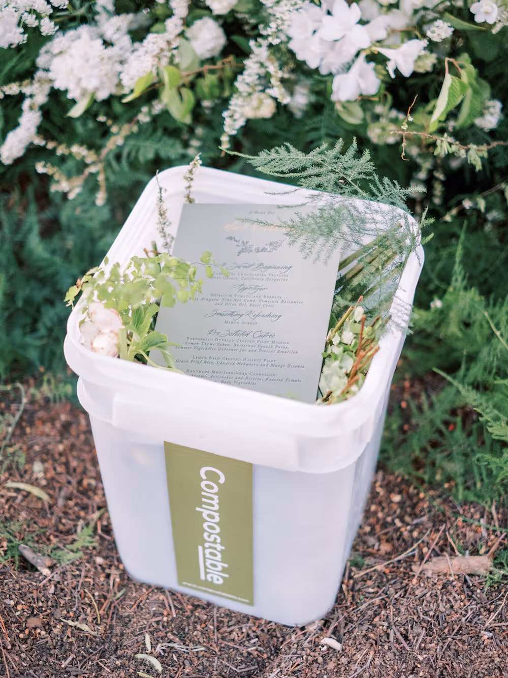 Photo of florals and wedding invitation placed in a white compost bin.