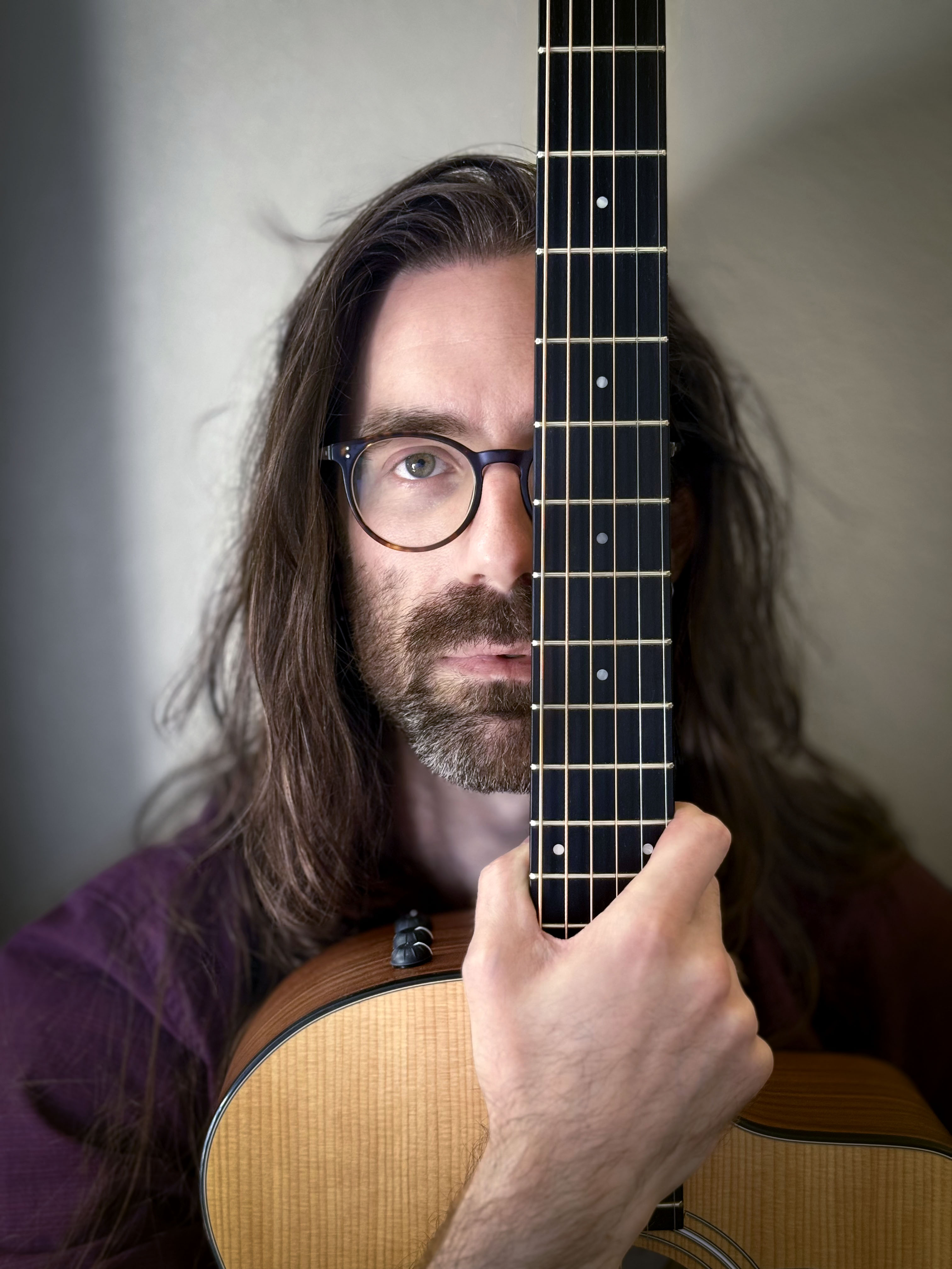 A headshot of a man with long hair and a short beard, wearing glasses, looking directly at the viewer while covering his left eye with the fretboard of a guitar held vertically in his right hand.