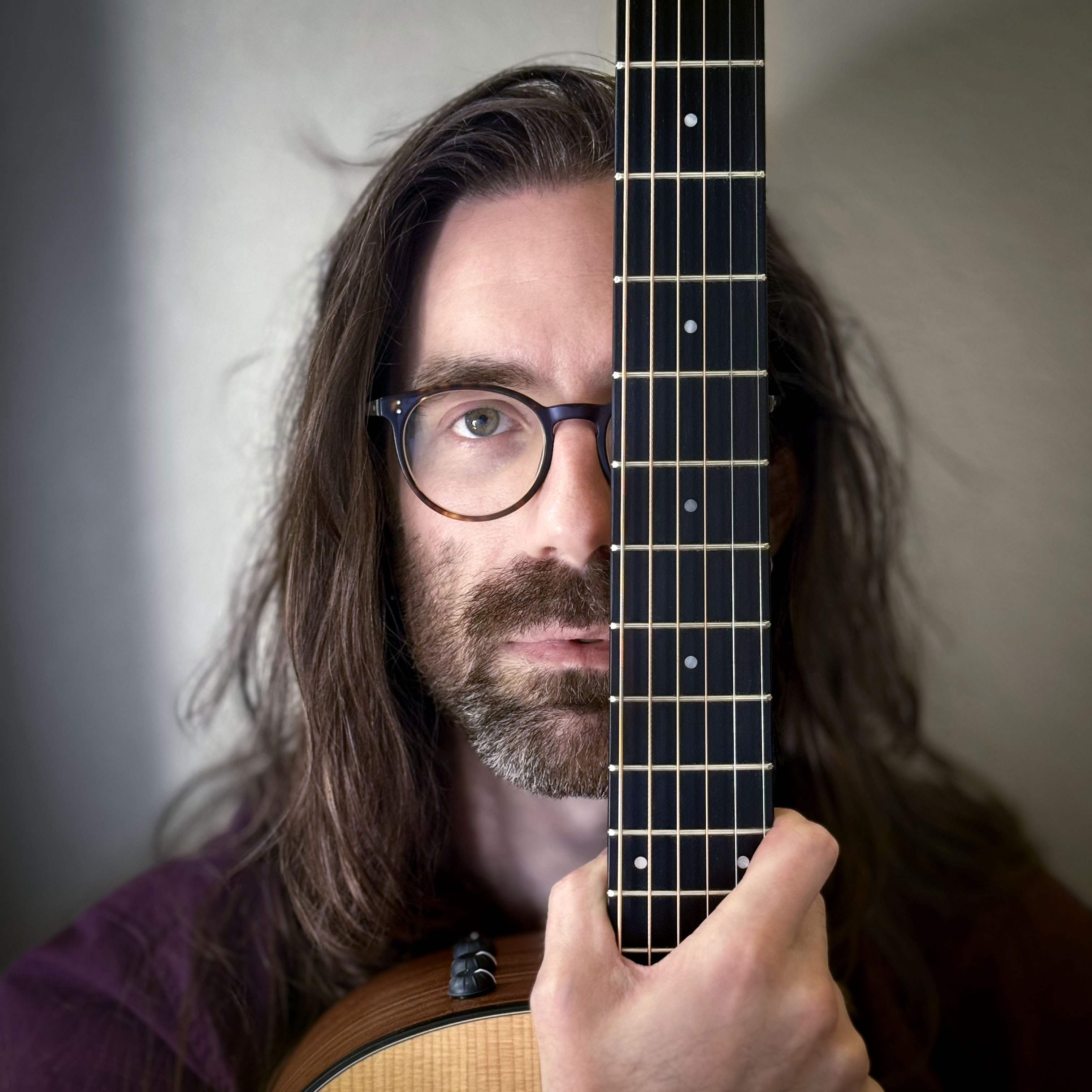 A headshot of a man with long hair and a short beard, wearing glasses, looking directly at the viewer while covering his left eye with the fretboard of a guitar held vertically in his right hand.