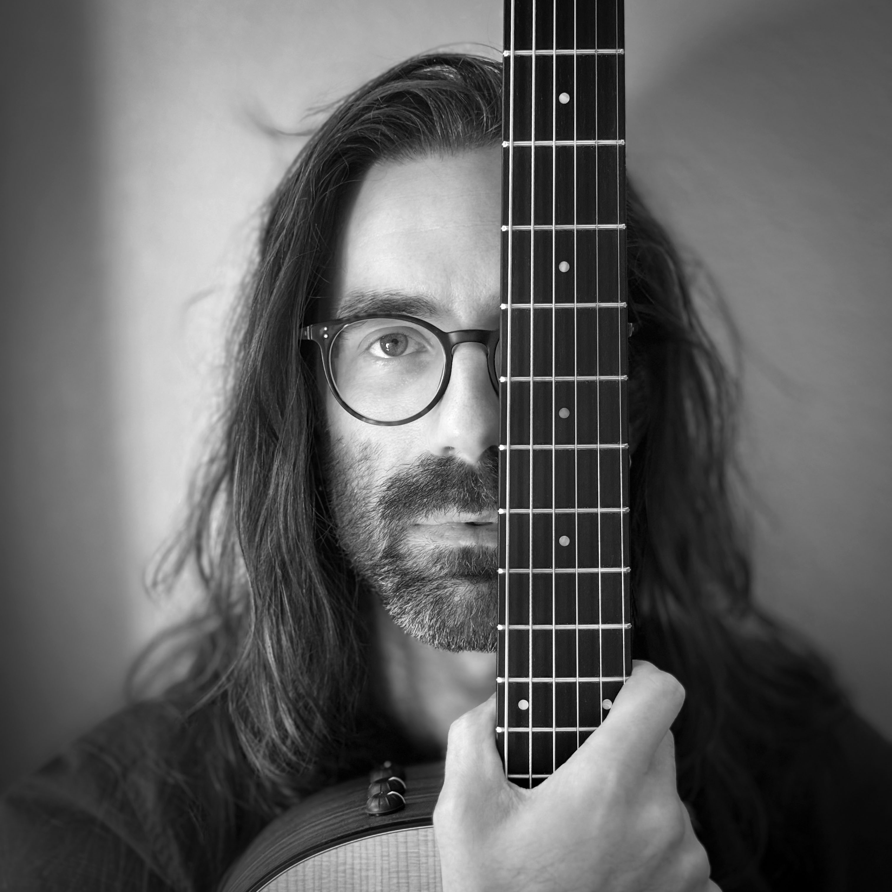 A grayscale headshot of a man with long hair and a short beard, wearing glasses, looking directly at the viewer while covering his left eye with the fretboard of a guitar held vertically in his right hand.