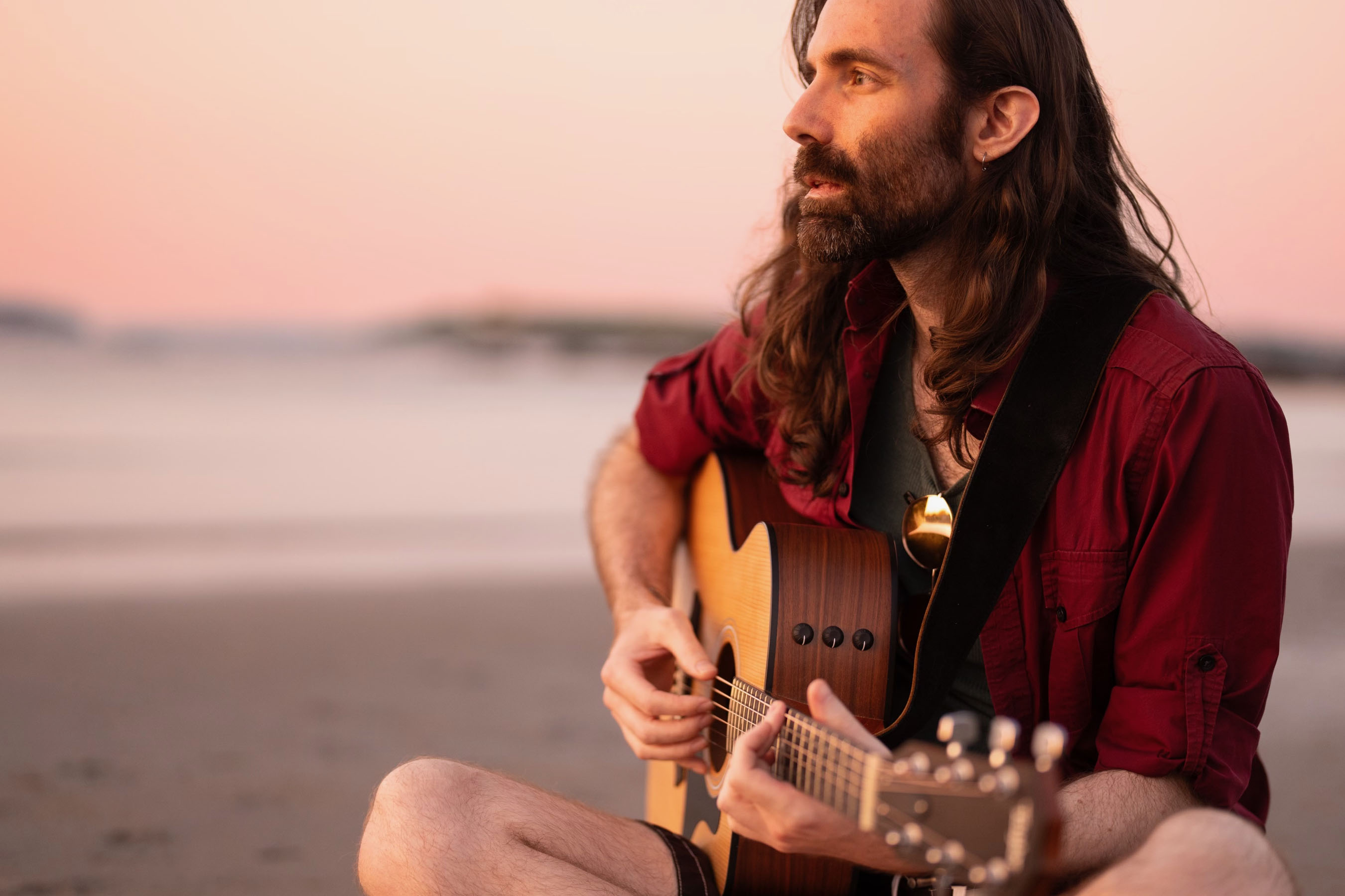 A man with long hair and a short beard plays a steel-string acoustic guitar while sitting on a sandy beach, looking toward the water.