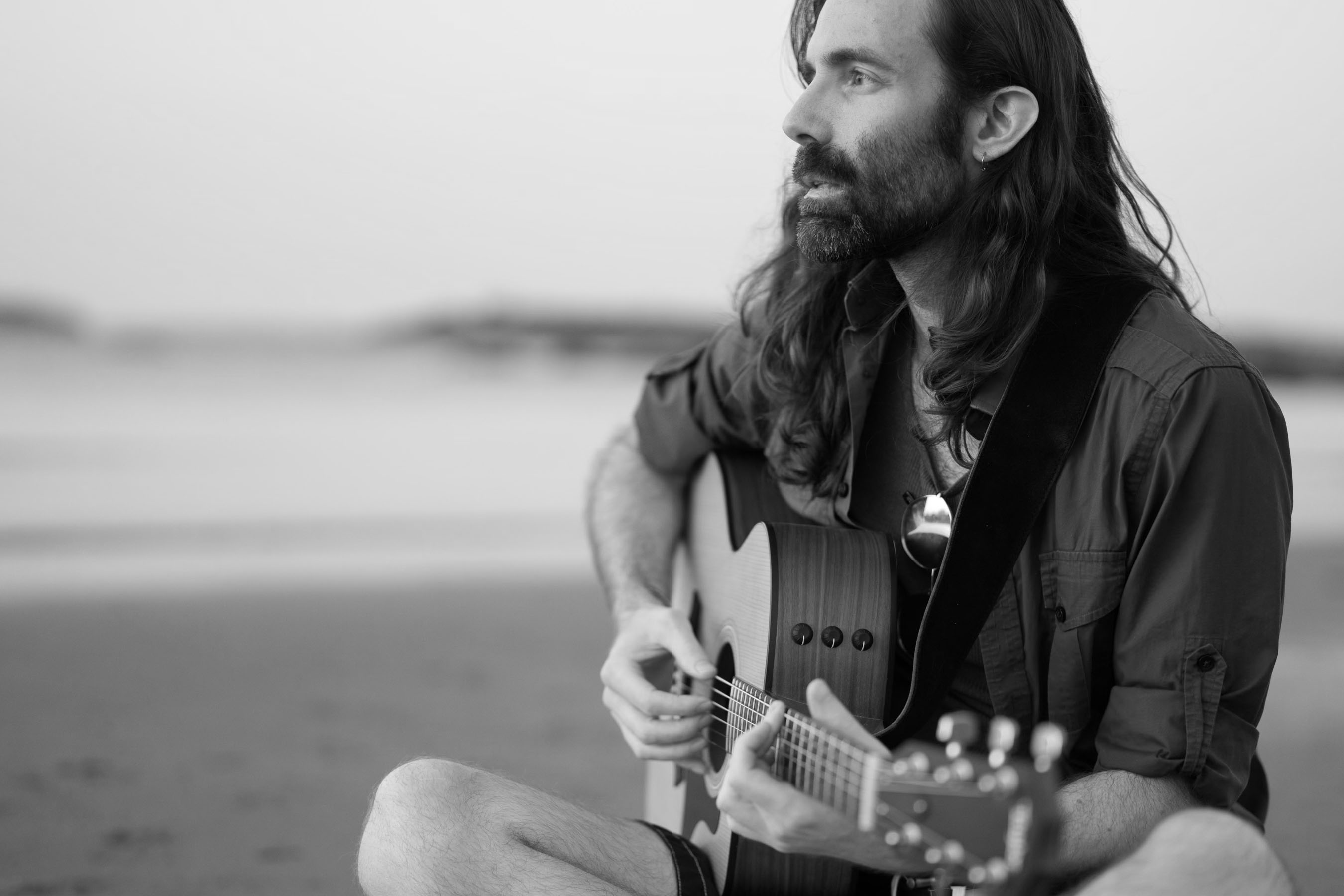 A grayscale photo of a man with long hair and a short beard plays a steel-string acoustic guitar while sitting on a sandy beach, looking toward the water.