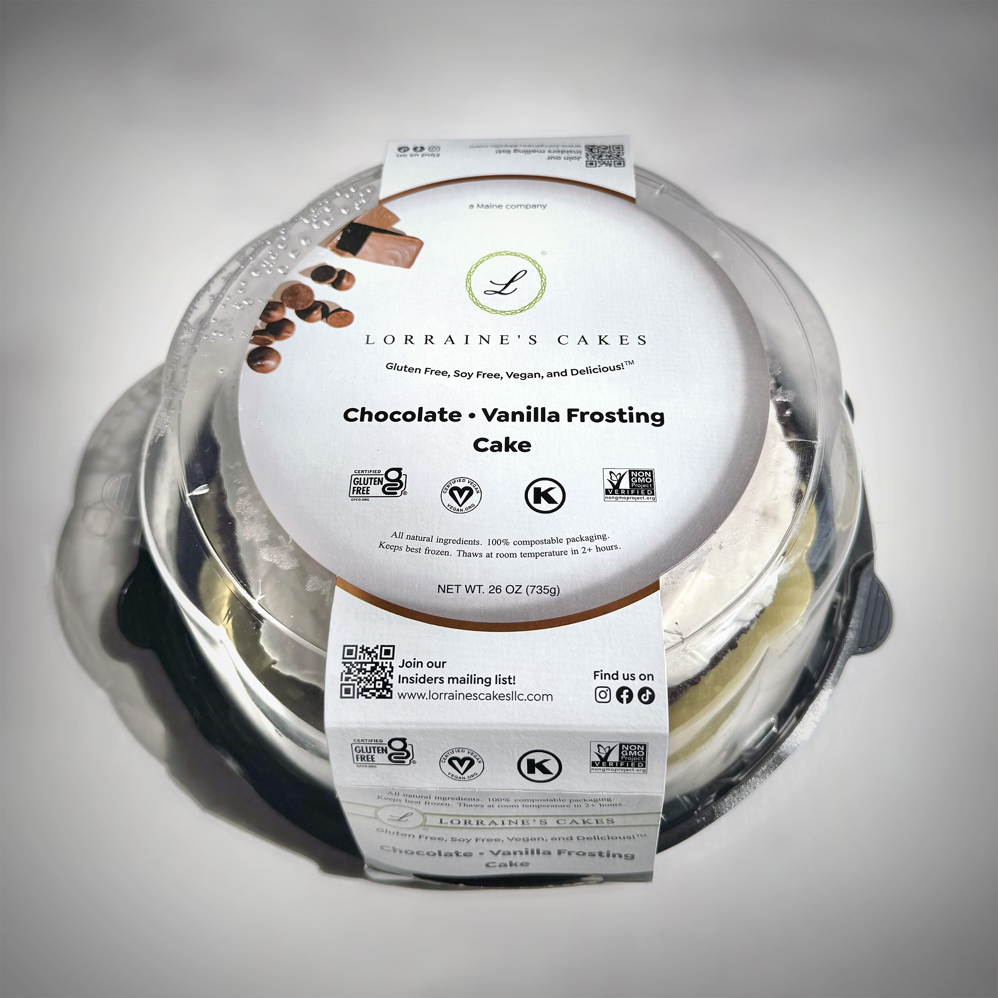 A three-quarter–angle view of a Lorraine's Cakes chocolate cake in its package: a thick card-stock sleeve printed with branded graphics around a clear, compostable plastic dome.