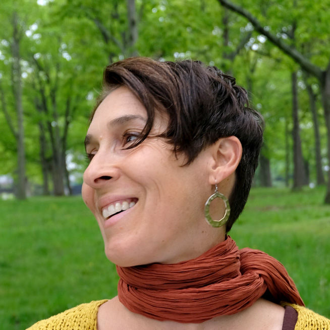 A smiling woman with short, dark brown hair wearing a terracotta-toned scarf and yellow knit shirt, looks to the left standing in lush green woods