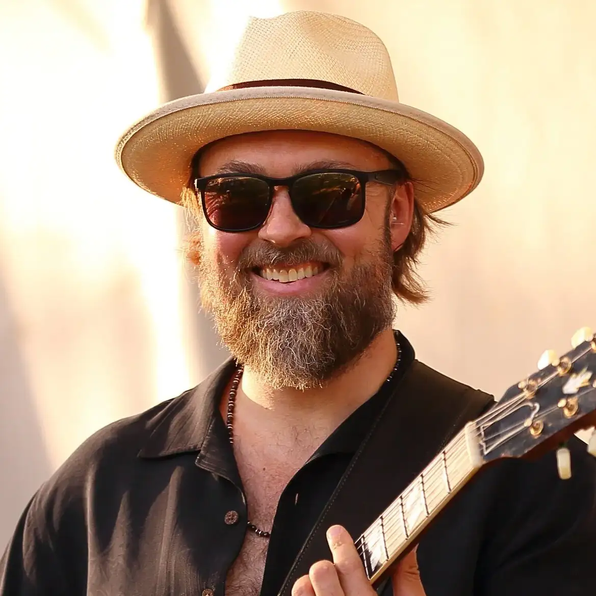 Bearded man wearing a light straw hat and dark sunglasses smiling while holding a guitar, dressed in a black shirt with a beaded necklace, before a sunlight stage backdrop.