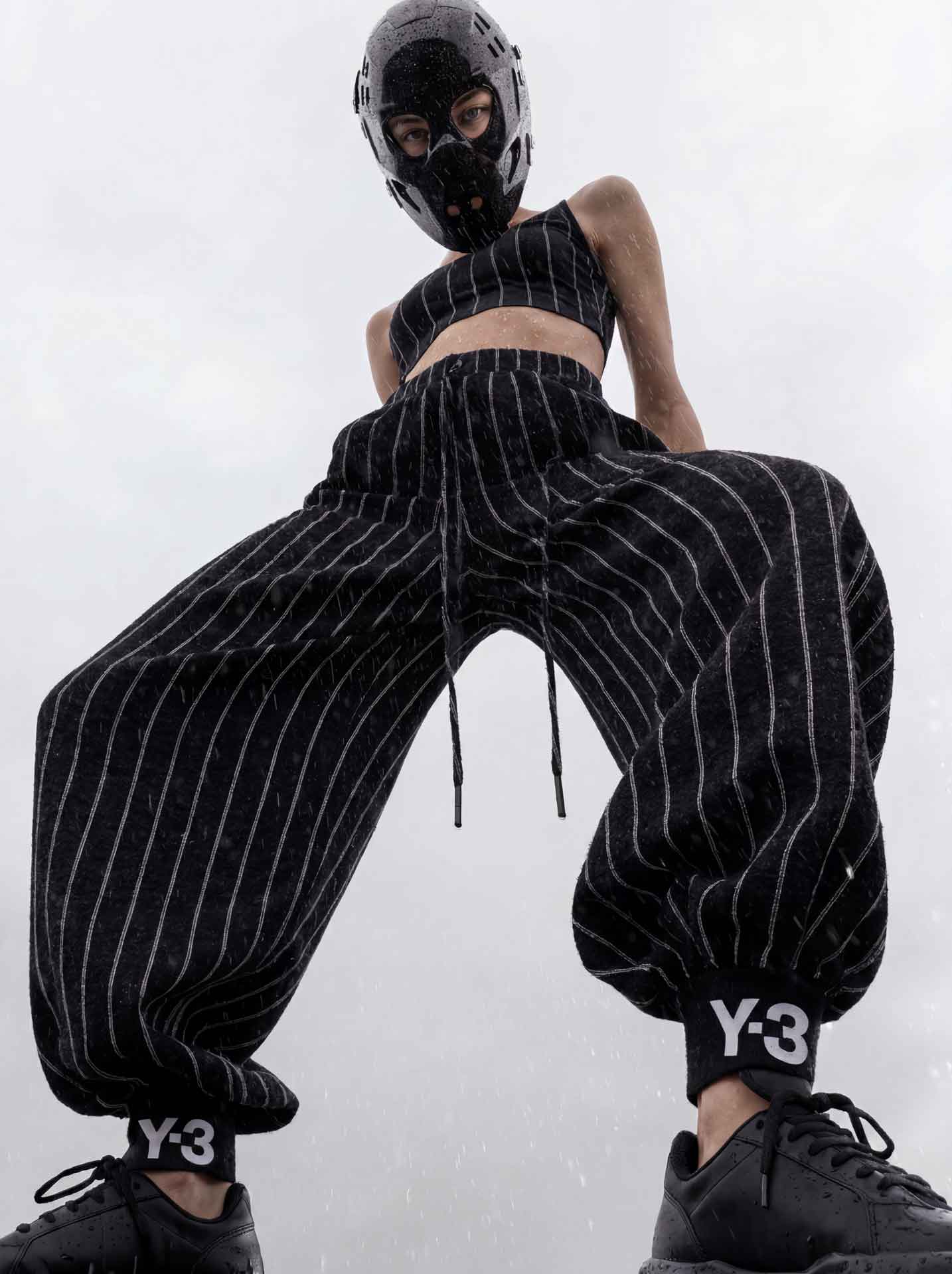 Low-angle view of a person wearing a black pinstriped outfit with Y-3 branding on the pants cuffs and a futuristic black helmet.