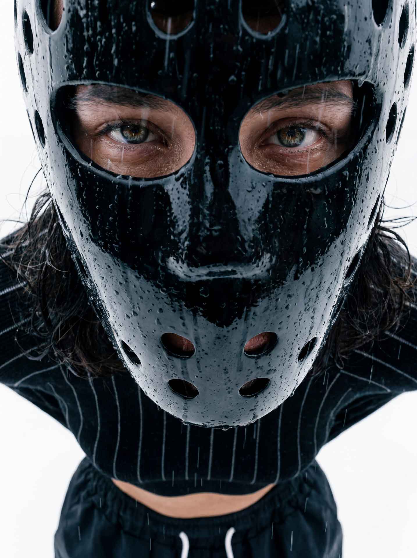 Person wearing a water-covered black mask with holes, looking directly at the camera in the rain.