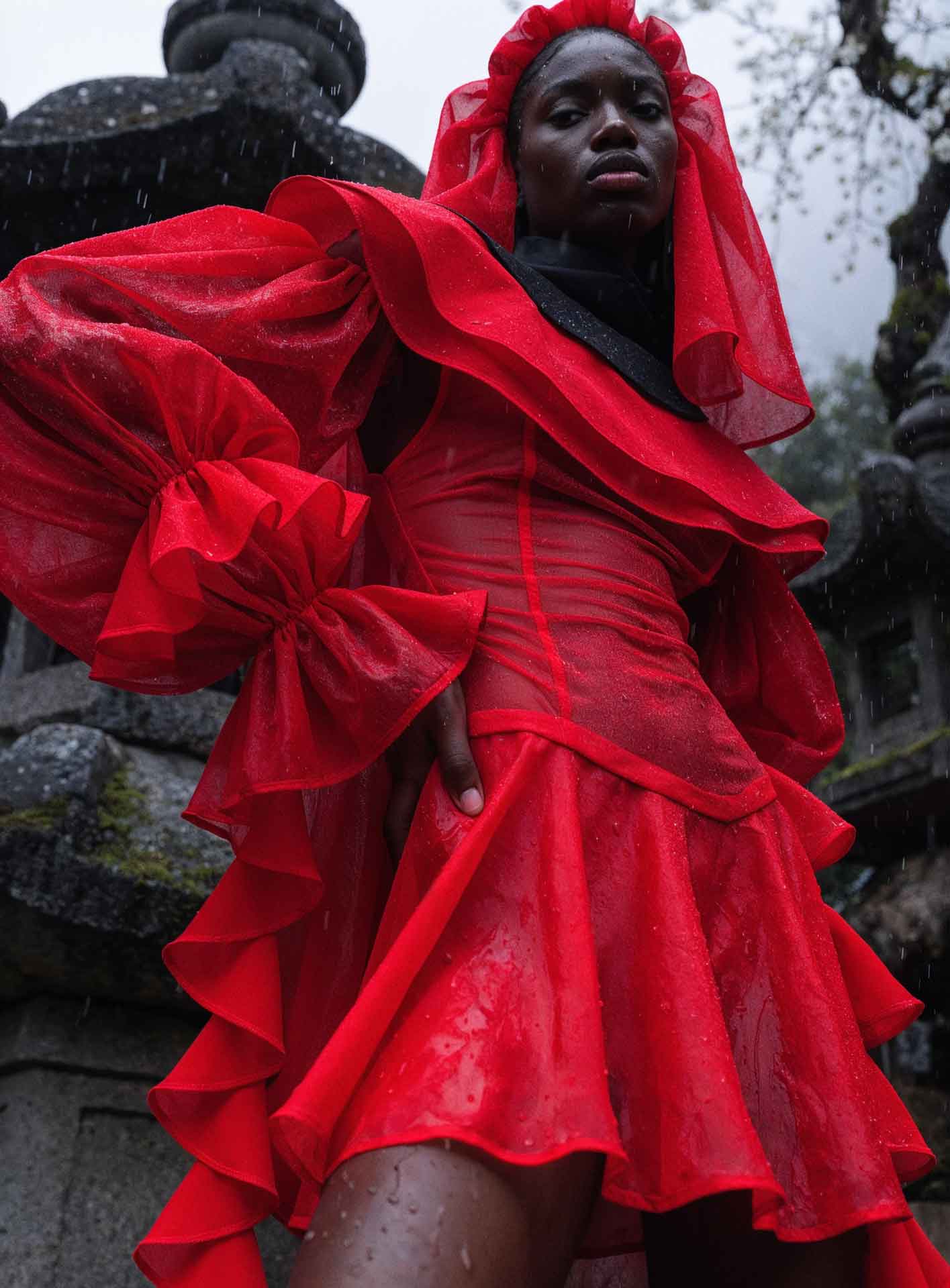 Person wearing a vibrant, ruffled red dress with puffed sleeves and a hood, standing outdoors in the rain near stone structures.