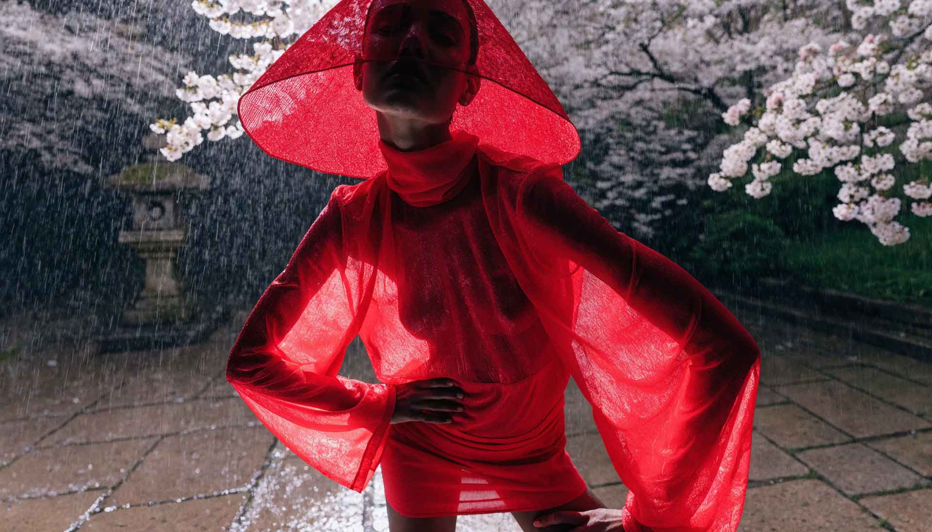 Person wearing a sheer red dress and wide red hat, posing with hands on hips in the rain near cherry blossoms.