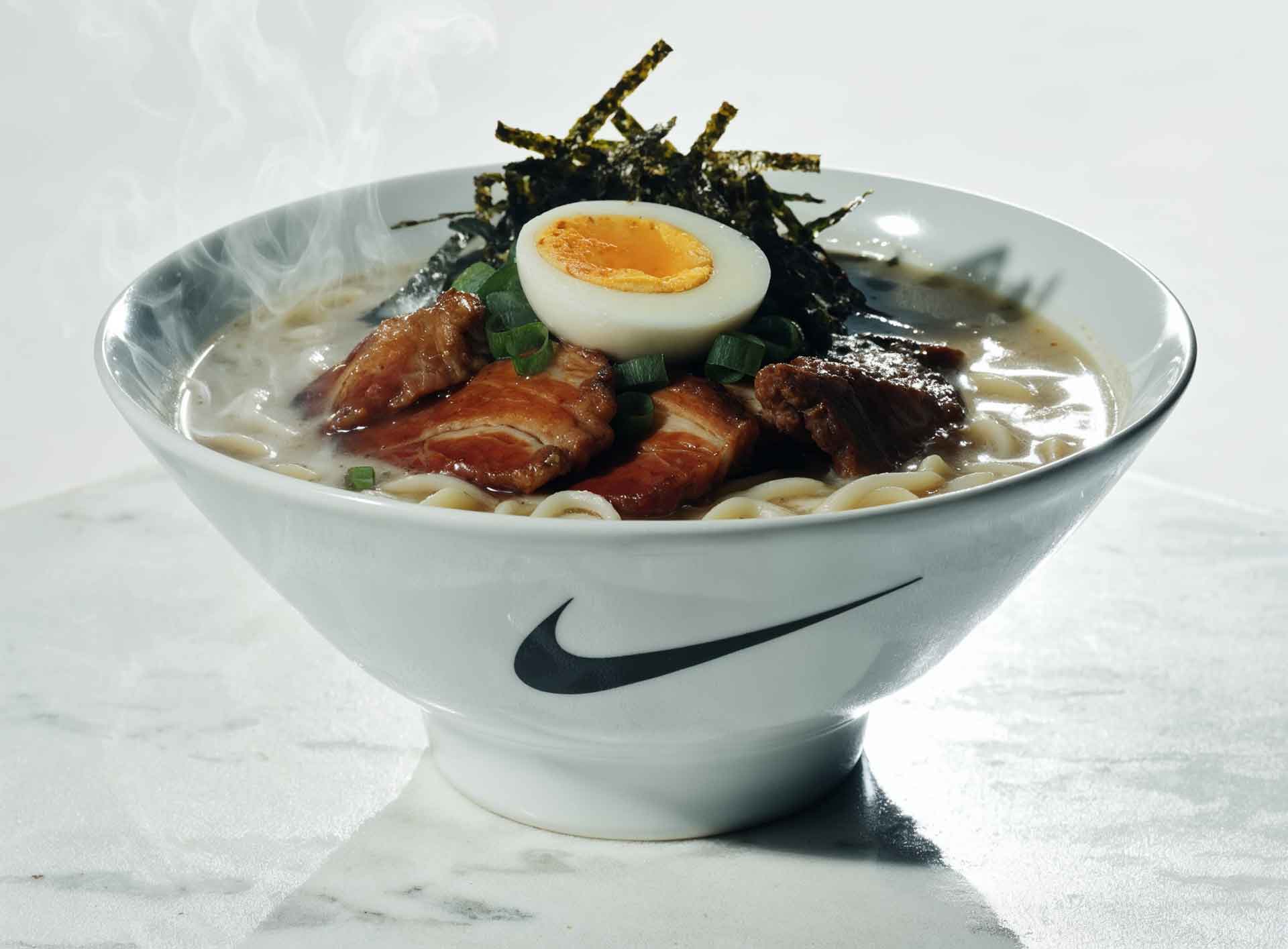 Steaming bowl of ramen with sliced pork, half a boiled egg, green onions, and seaweed strips in a white bowl with a black Nike swoosh logo.