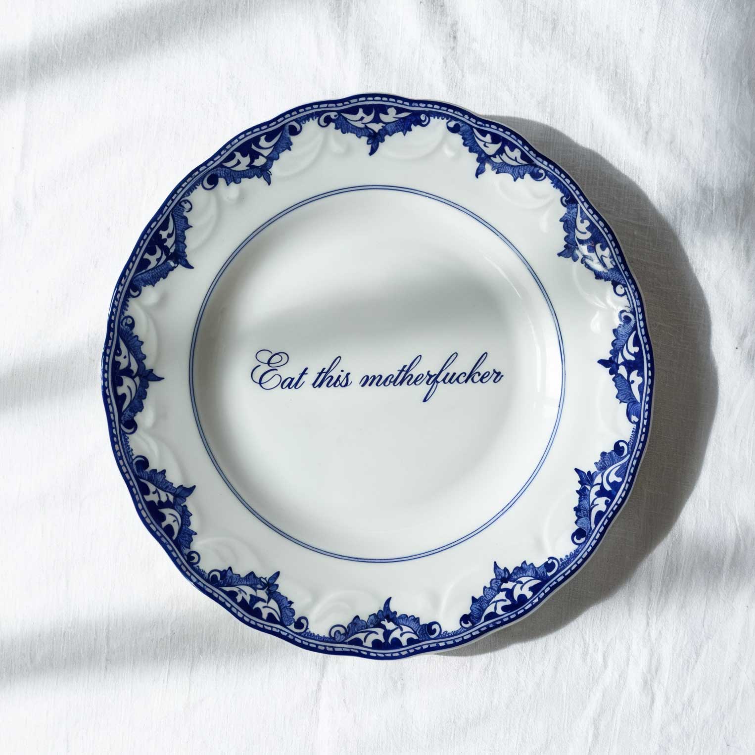White ceramic plate with blue ornate border and the phrase 'Eat this motherfucker' in cursive script at the center.