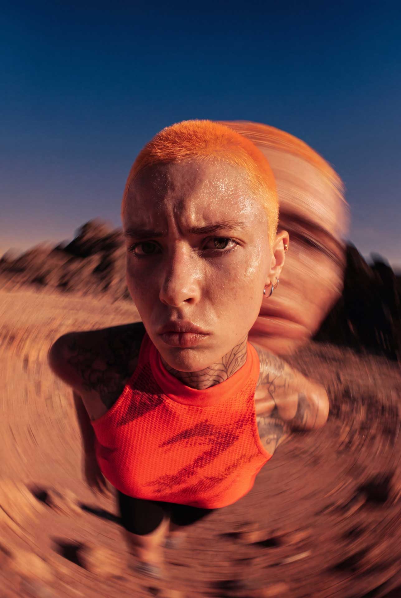 Person with short bright orange hair and tattoos wearing an orange sleeveless top, looking intensely at the camera with a motion blur effect around them in a desert setting.
