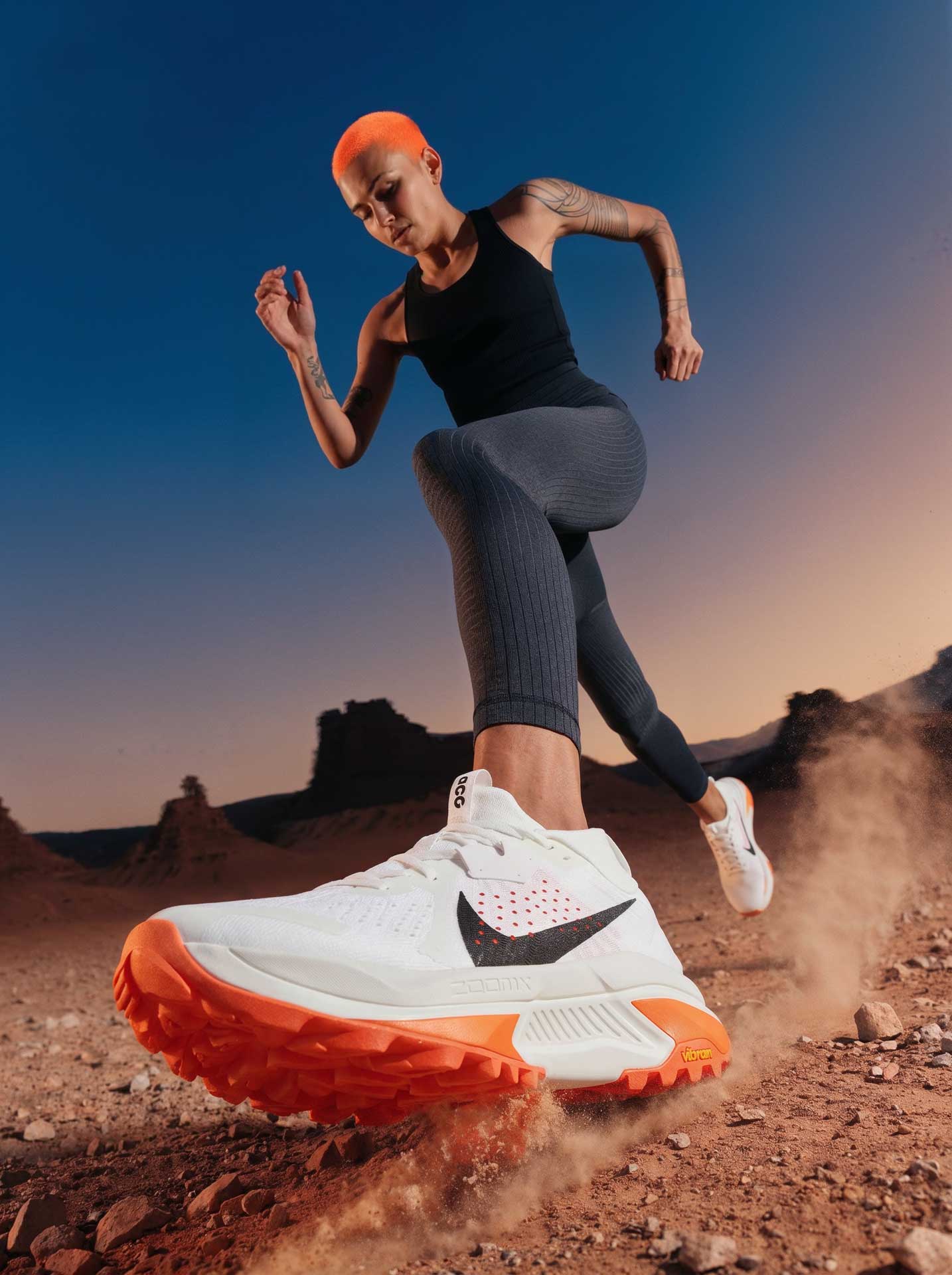 Person with bright orange hair running on rocky terrain wearing white and orange Nike running shoes, black top, and gray leggings.