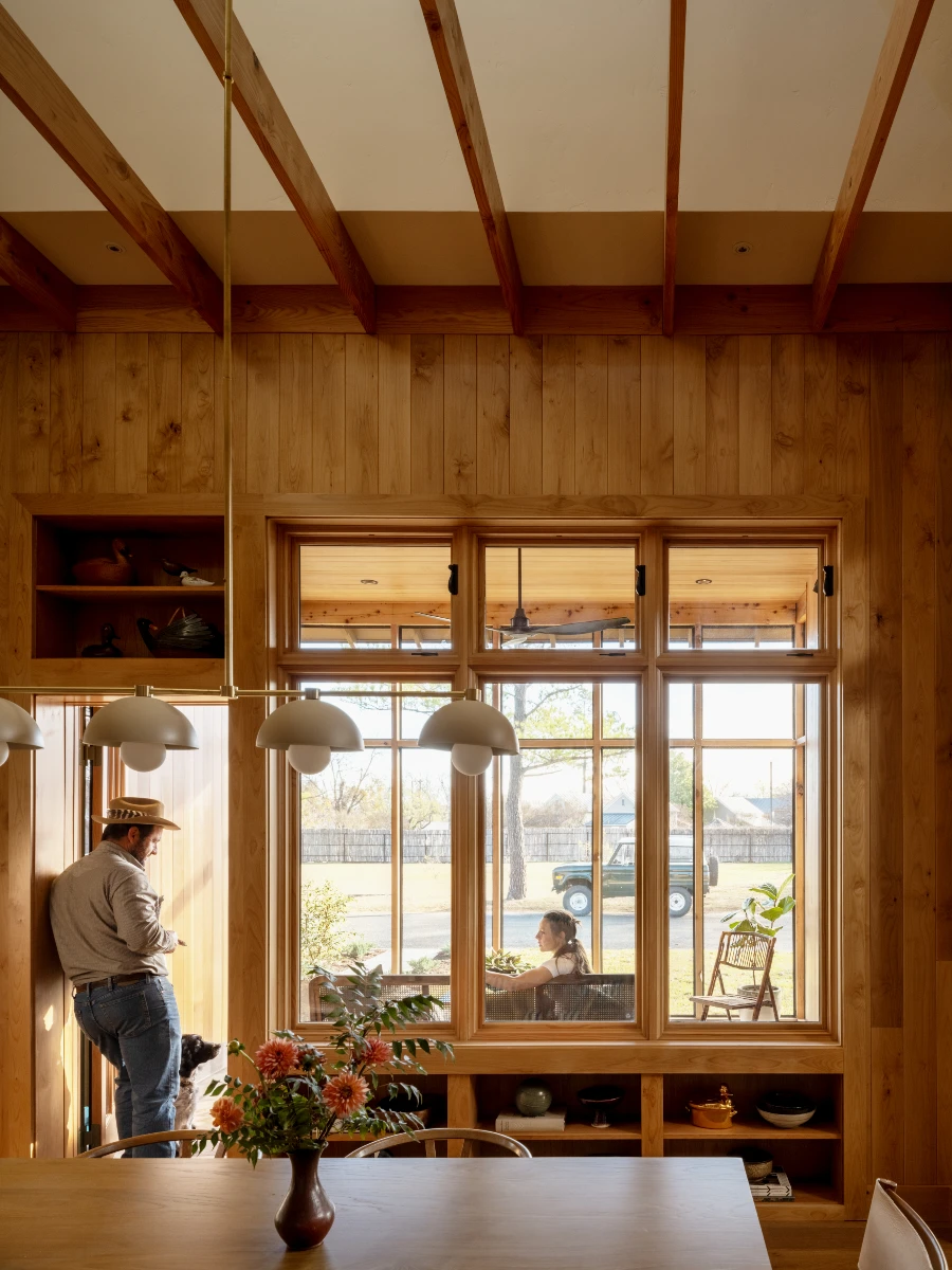 A shot of the dining room with man and dog standing in doorway, woman sitting in sunroom beyond