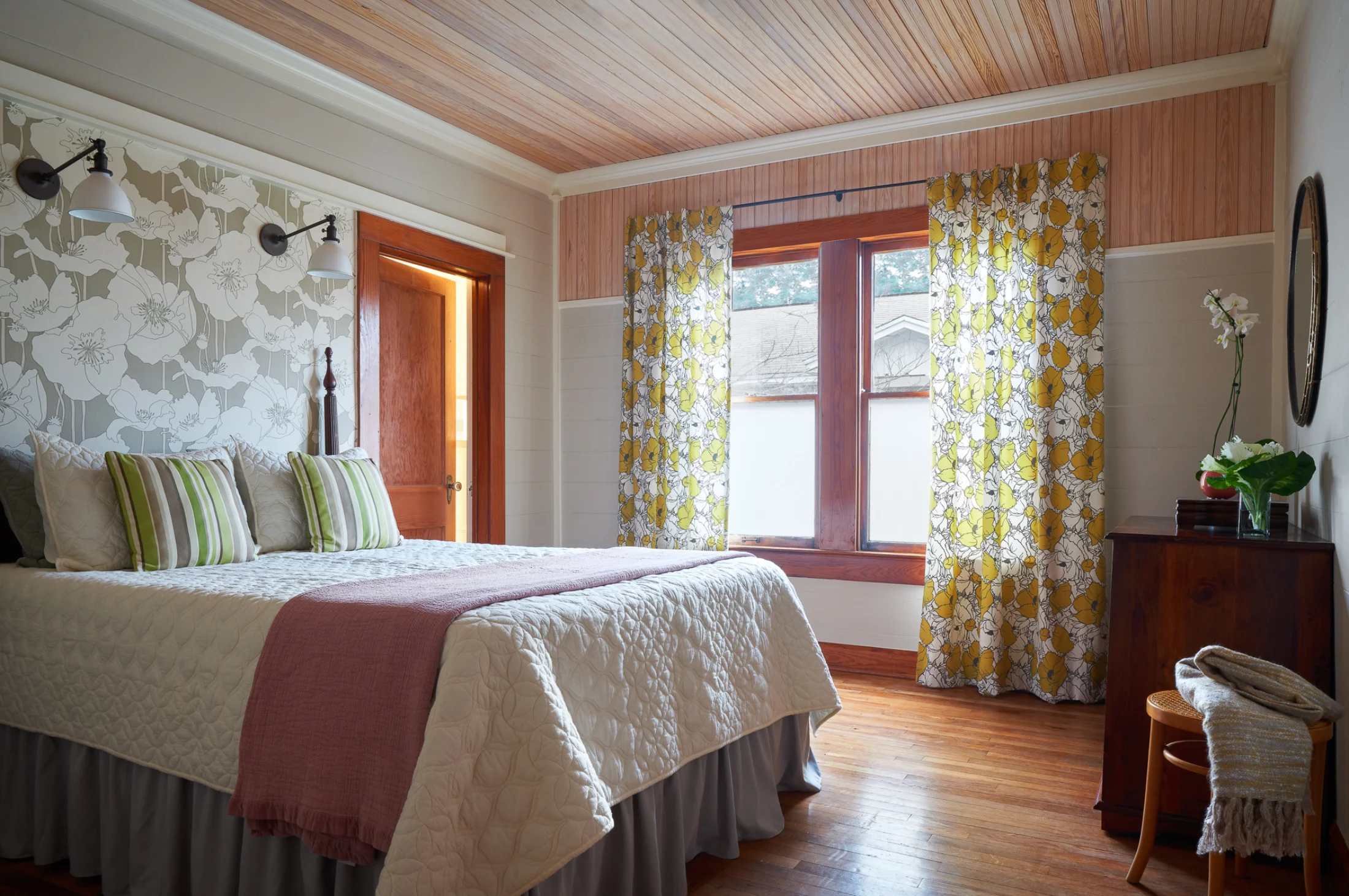 A Wende Bungalow bedroom furnished with a cozy vintage bed & dresser. The walls feature a neutral-toned floral wallpaper combined with wood paneling.