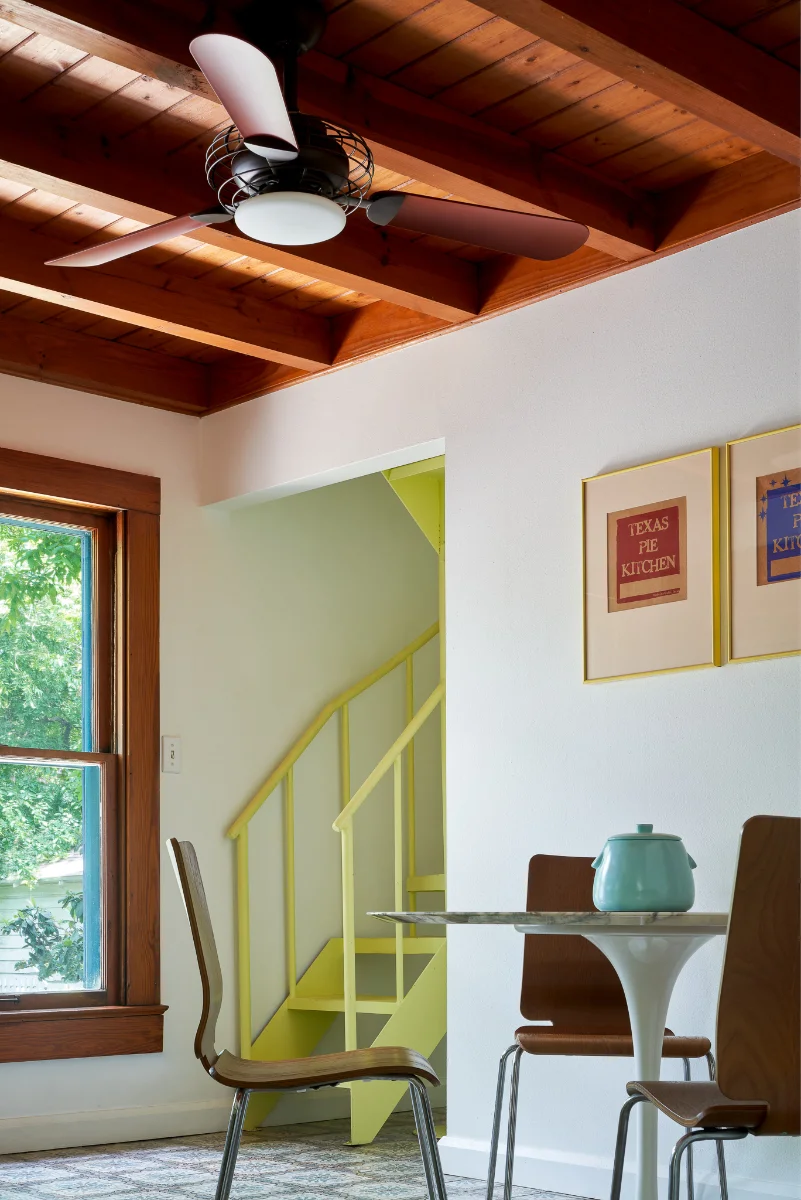 A dining area in Wende Bungalow appears is decorated with a light blue urn. The background shows a lemon yellow staircase near a window.