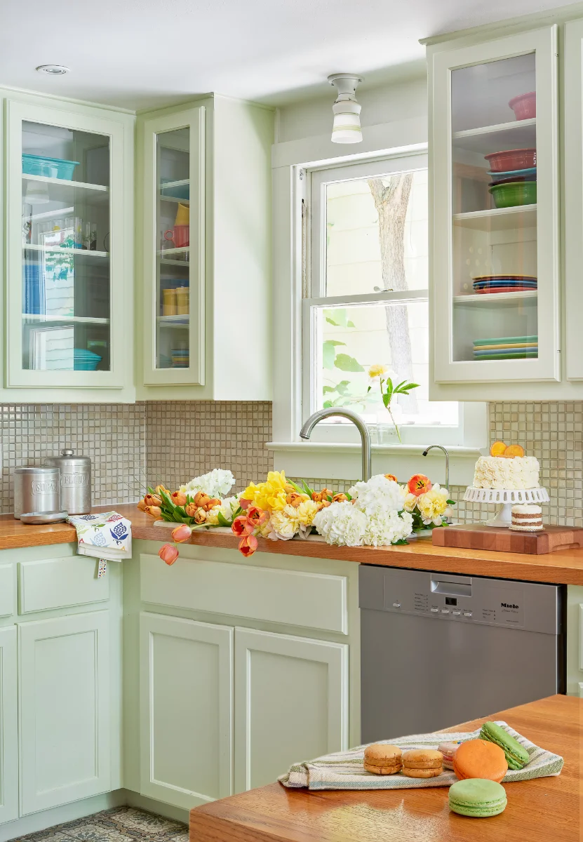 The kitchen of Wende Bungalow features pale green cabinetry and warm wood countertops. In the center, a sink is full of warm-hued flowers.