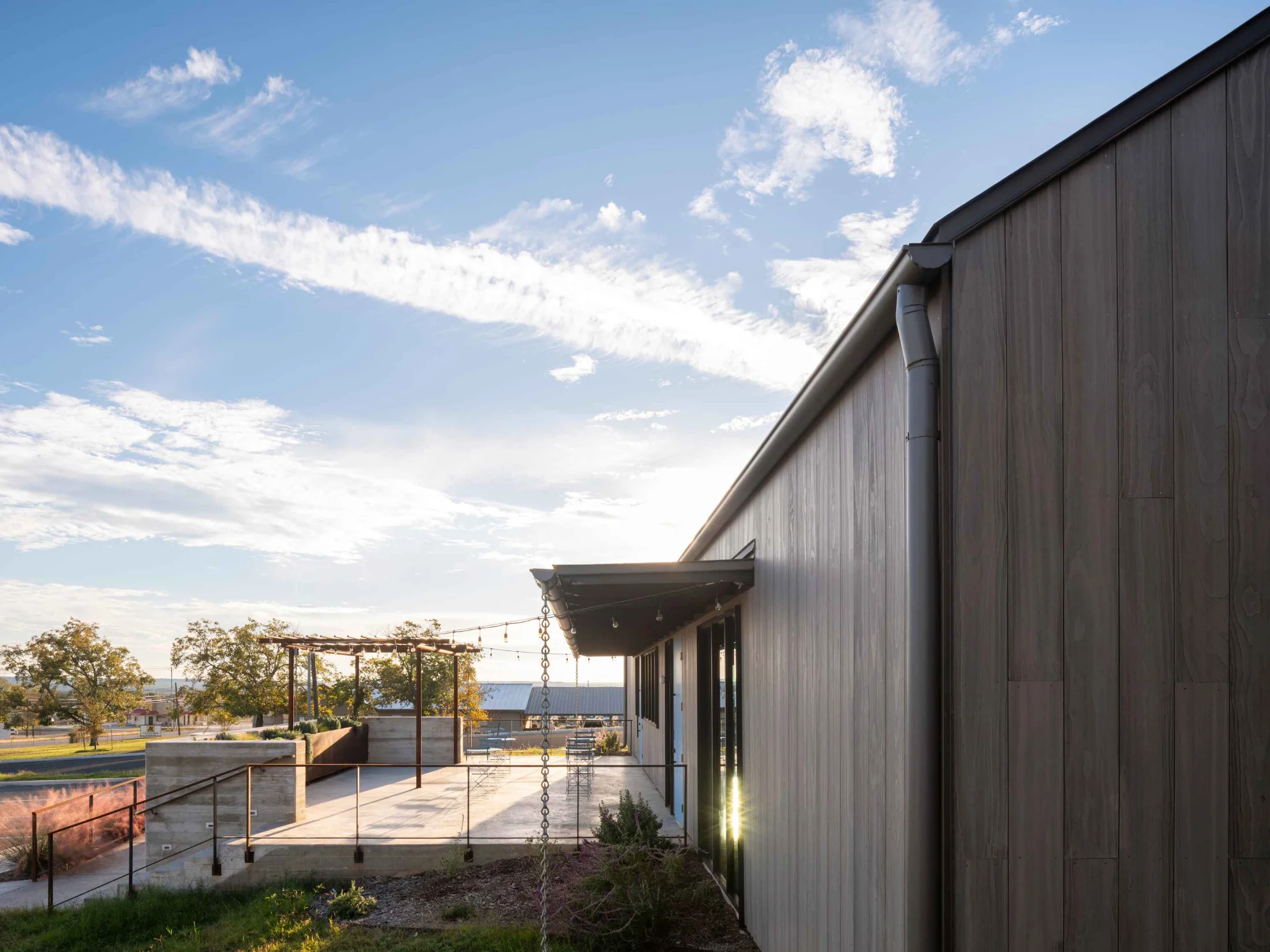A welcoming outdoor space landscaped with brushy plants & pergola. To the right, an exterior entrance to the FBG Office. An expansive sky is overhead.