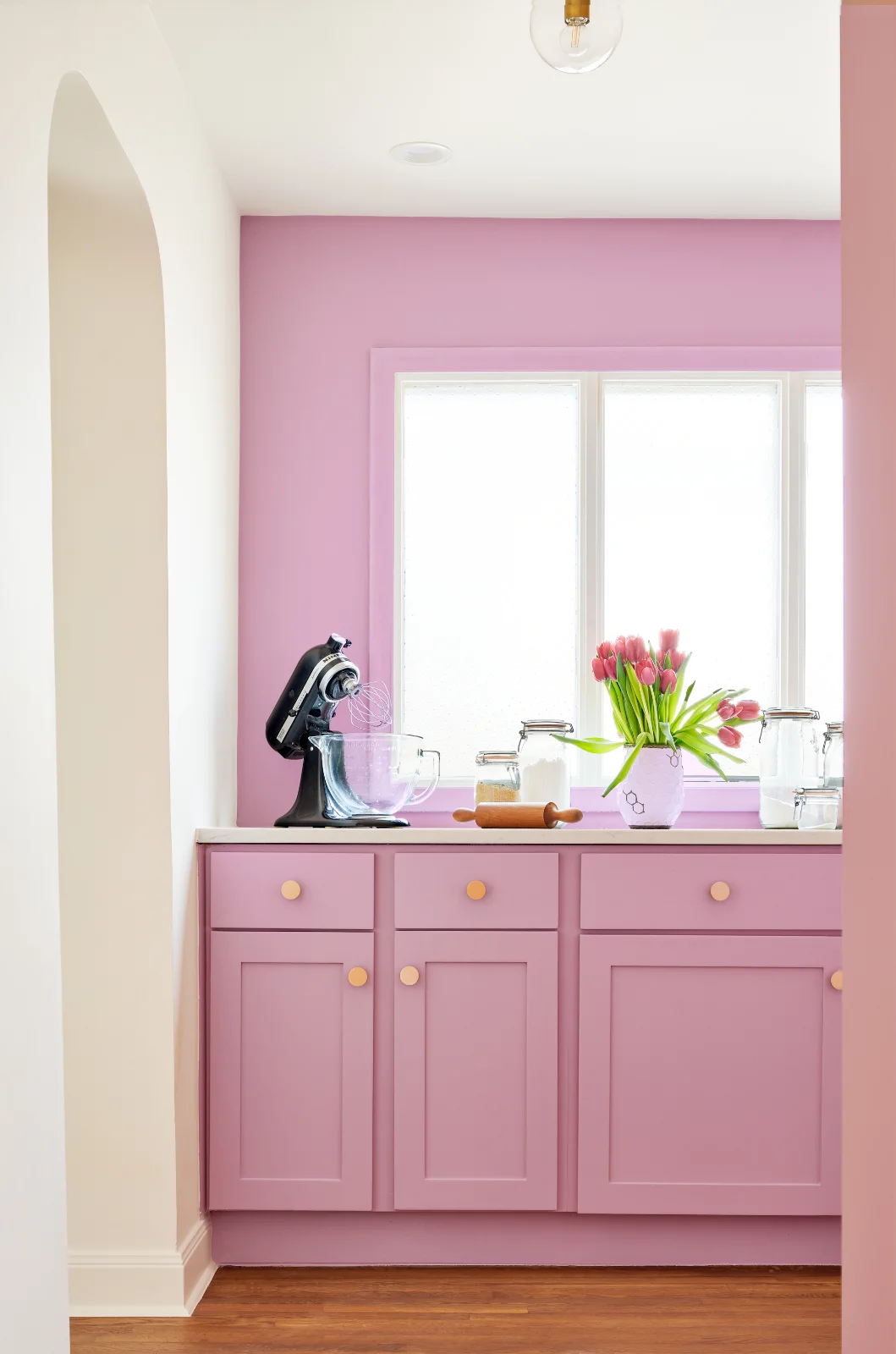 An area of the Queen Anne House is decorated with lilac pink cabinetry and walls. The counters hold baking supplies and a black KitchenAid mixer.