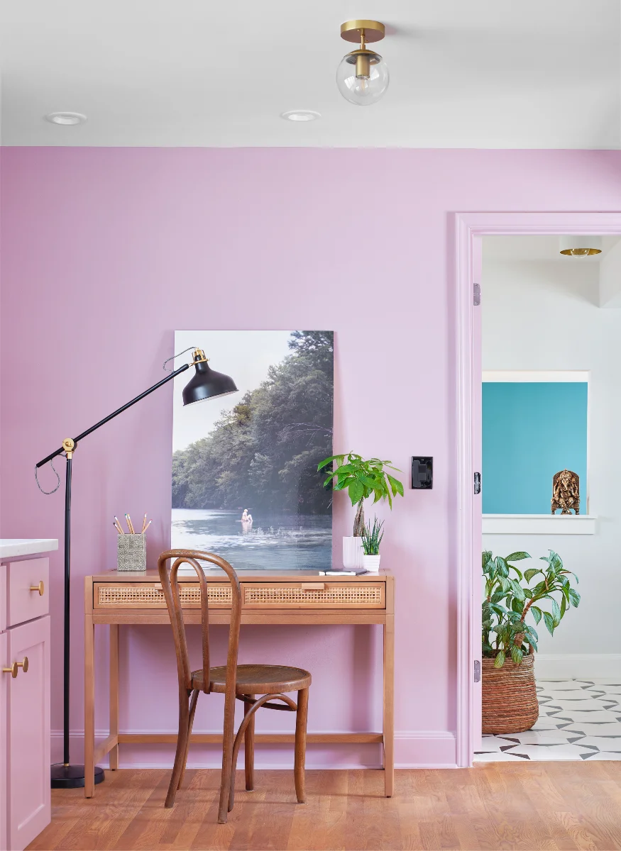 A room in the Queen Anne House is decorated with lilac pink walls and warm wood furniture. A desk and chair sit in the center of the image.