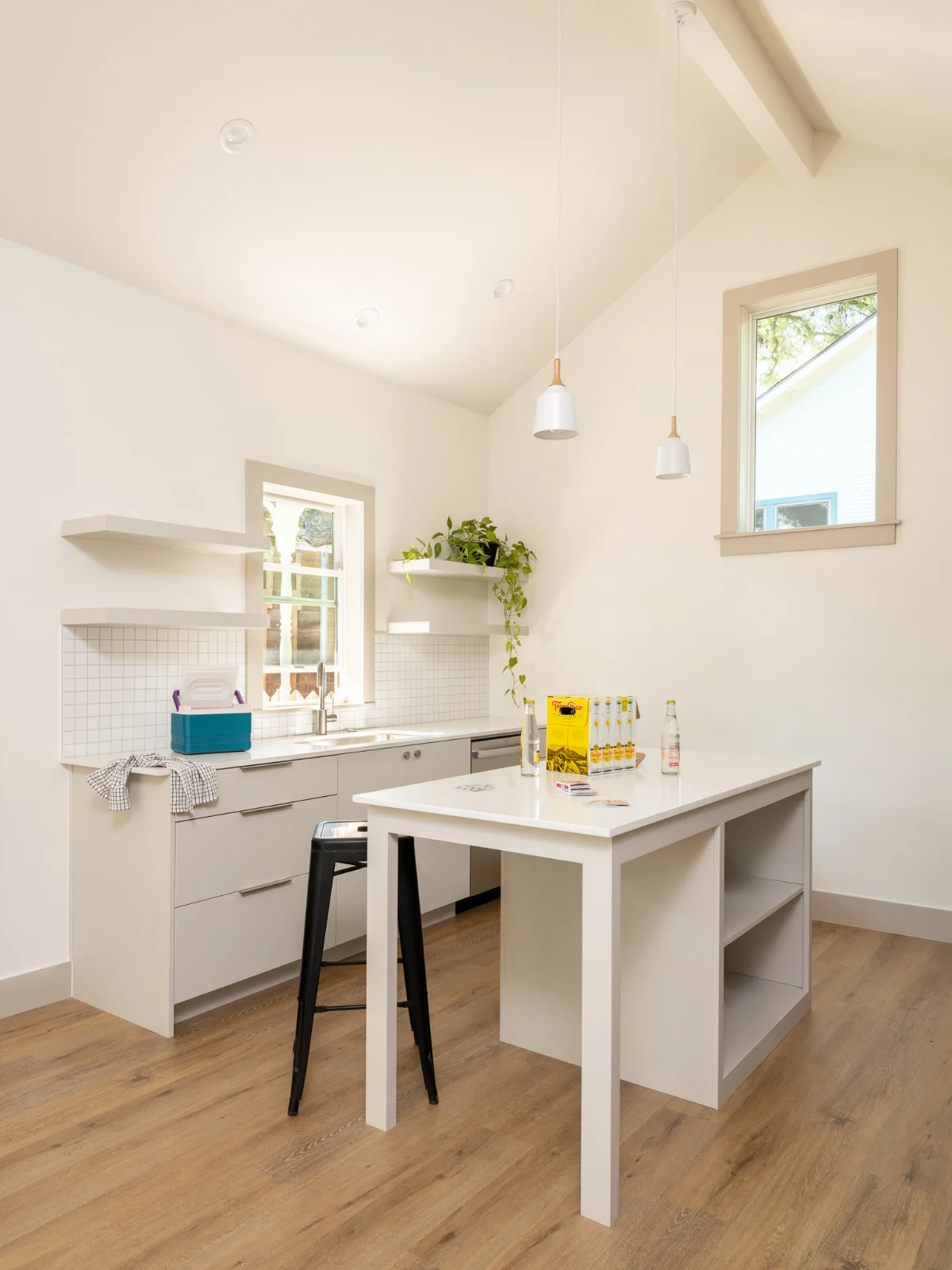 A corner of the Back House features a modern kitchen space with island seating. Cream cabinets are framed with a square white tile backsplash.