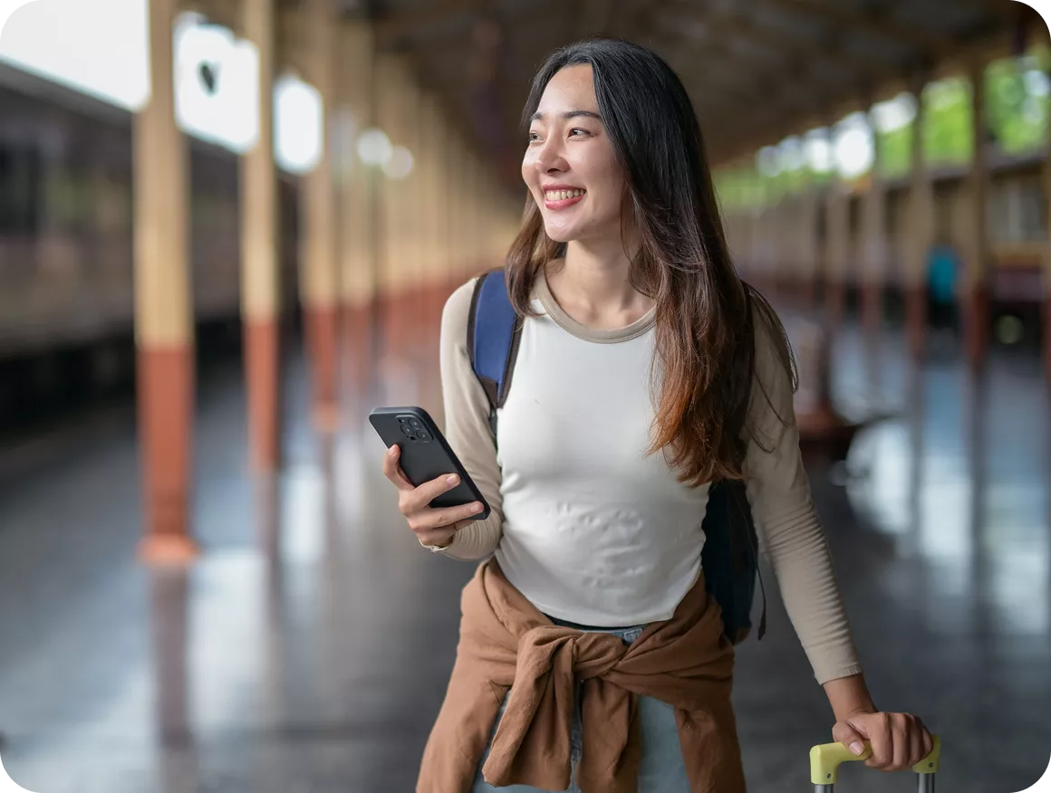 Smiling young woman holding a smartphone and luggage handle at a train station platform.