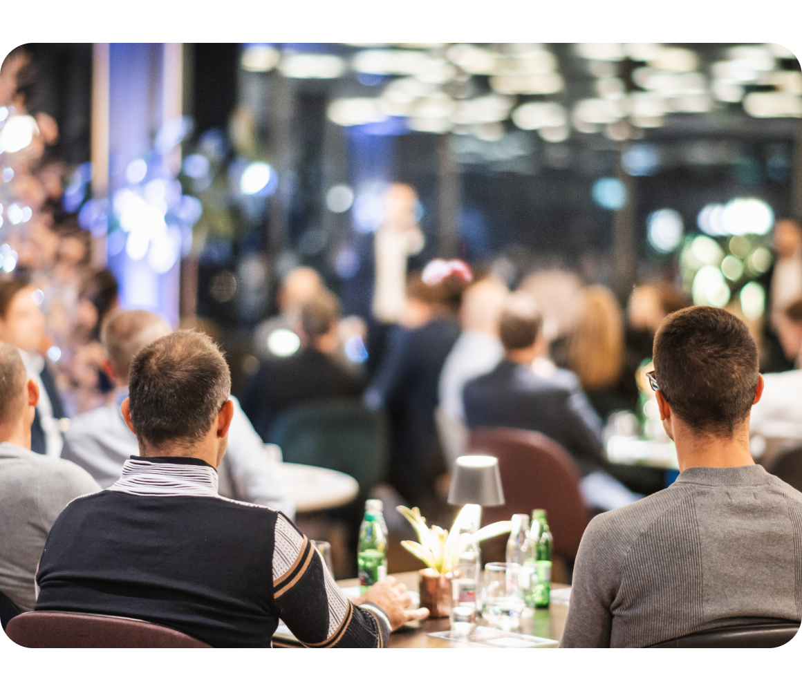 Audience seated at tables listening to a speaker in a softly lit conference room.