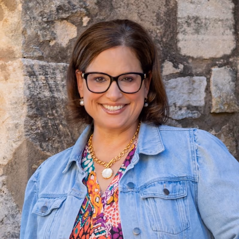 Smiling woman in denim jacket and colorful top, wearing glasses