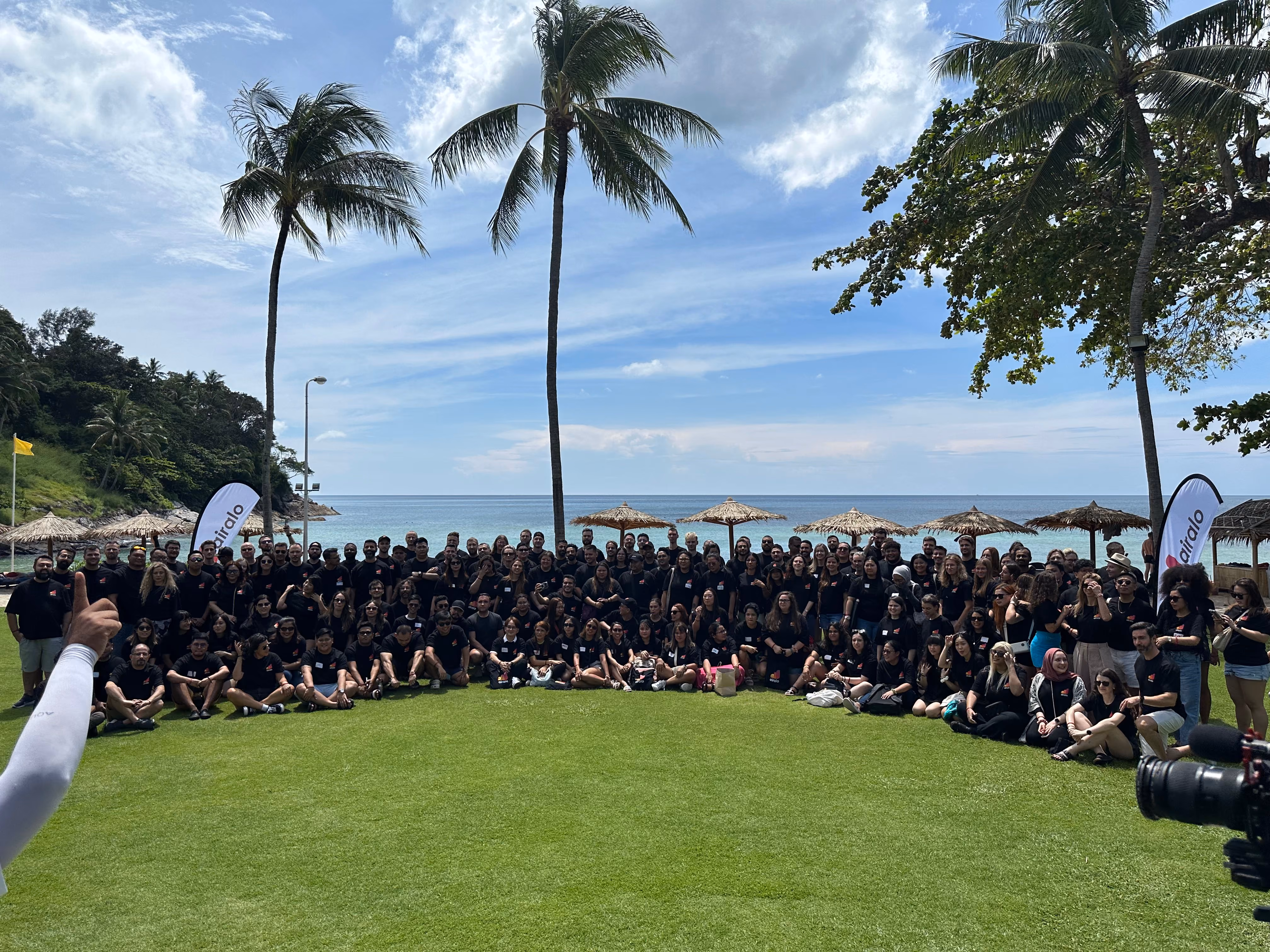 Large group of people wearing black shirts gathered on green grass near a beach with palm trees and thatched umbrellas under a partly cloudy sky.