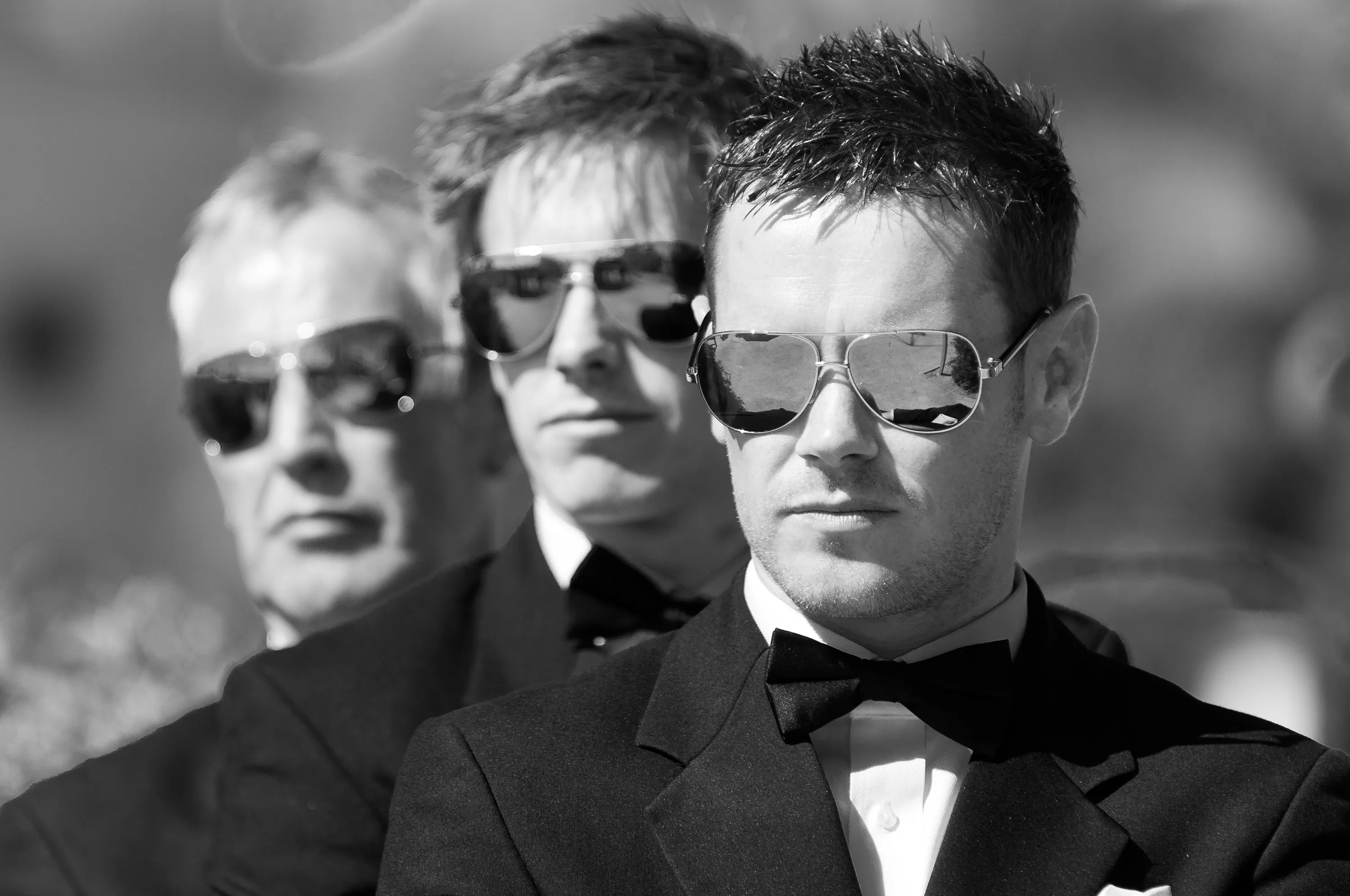 Three men in black tuxedos and bow ties wearing reflective aviator sunglasses, standing in a diagonal line outdoors.