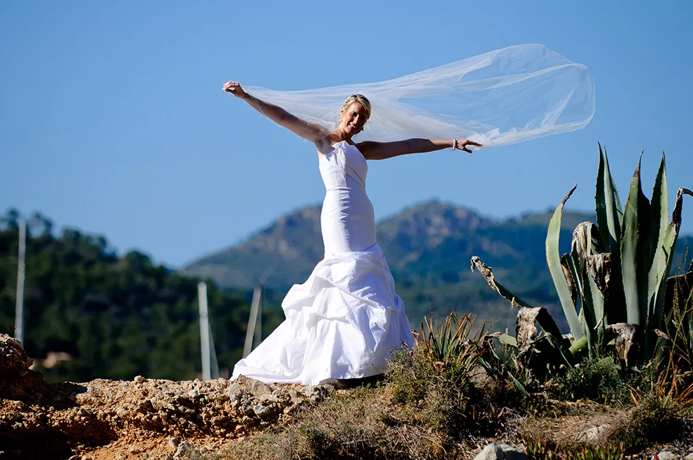 Bride in a white wedding gown and veil standing outdoors on rocky terrain with mountains in the background.