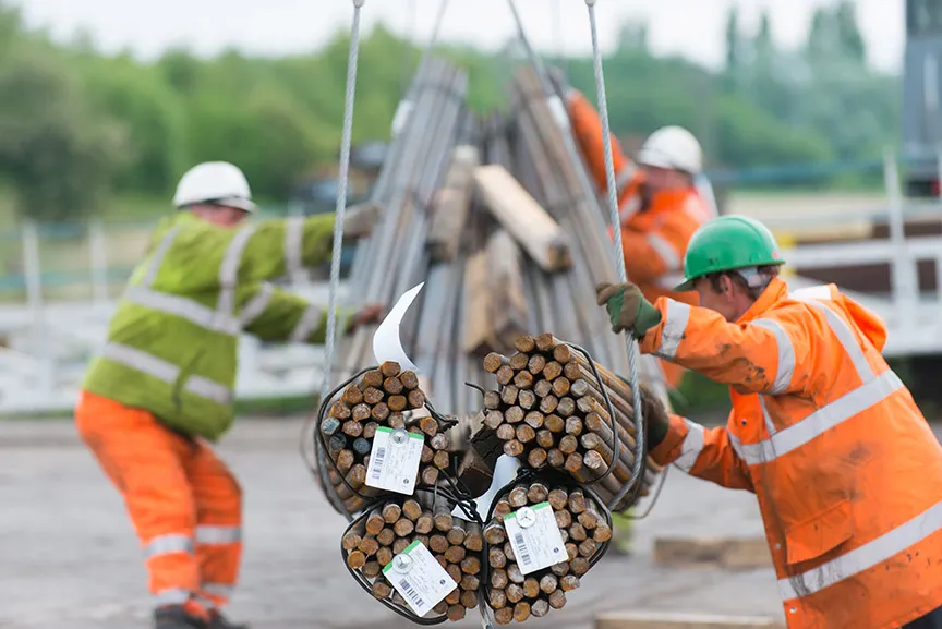 Three construction workers in safety gear guiding bundles of steel rods suspended by cables at a construction site.