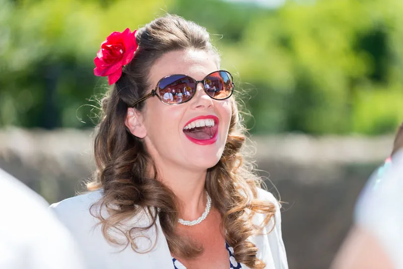 Woman with curly hair, red flower in hair, wearing sunglasses and a pearl necklace, laughing outdoors.