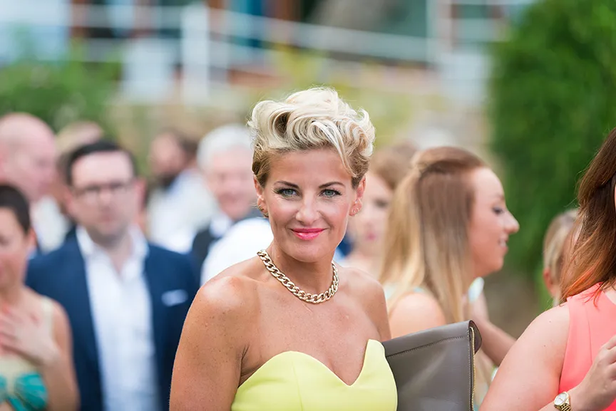 Smiling woman with short blonde hair wearing a yellow strapless dress and gold chain necklace at an outdoor event.