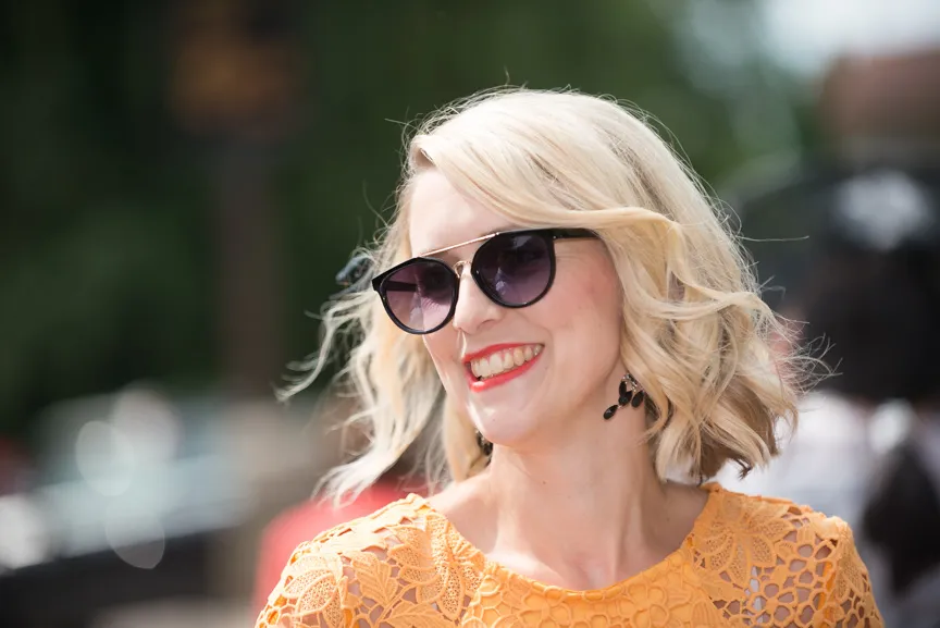 Smiling woman with short blonde hair wearing dark sunglasses and an orange lace top outdoors.