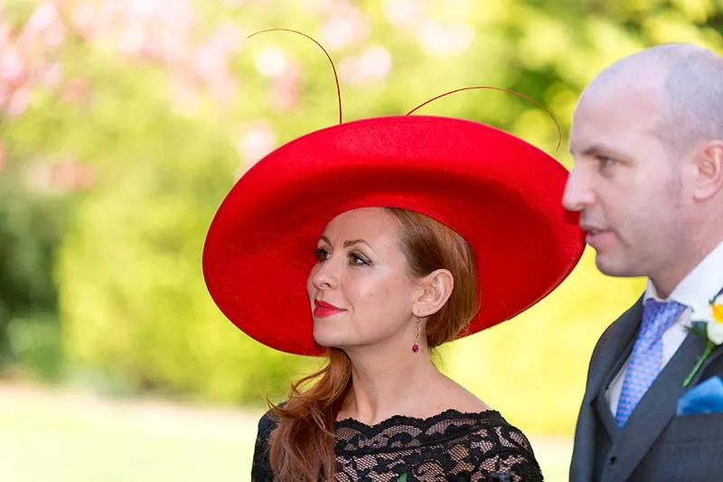 Woman wearing a large red hat and black lace dress standing next to a man in a suit with a blue tie and boutonniere.
