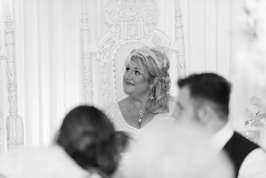 Smiling bride with a veil seated on an ornate white throne, looking slightly upward to the side.