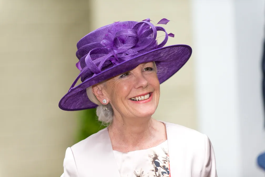 Smiling elderly woman wearing a large purple decorative hat and light-colored outfit.