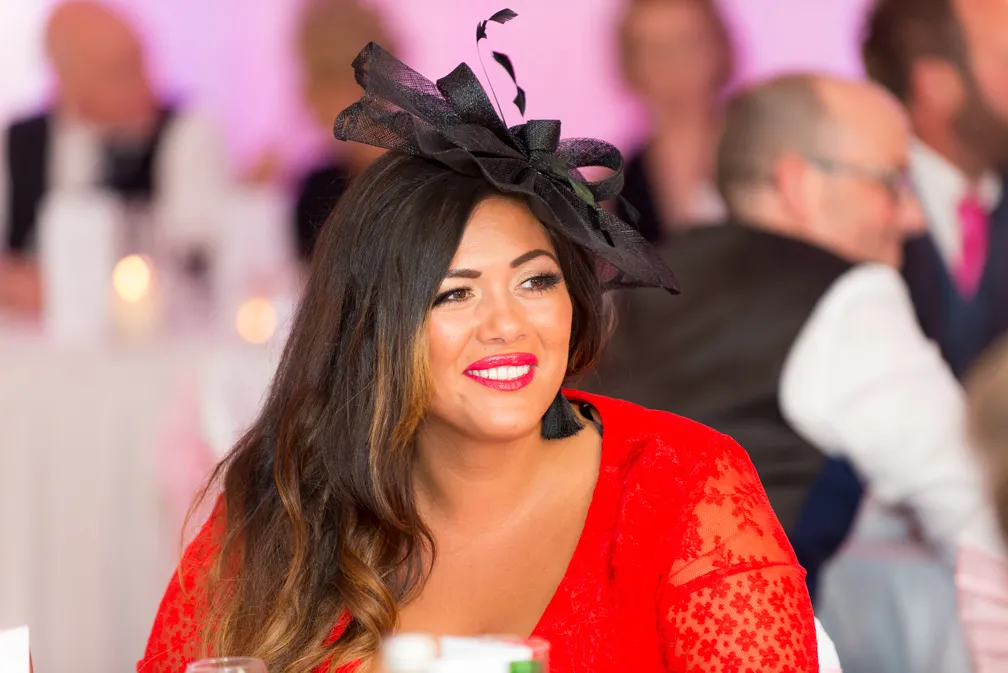 Woman with long dark hair wearing a black fascinator and bright red lace dress, smiling at a social event.