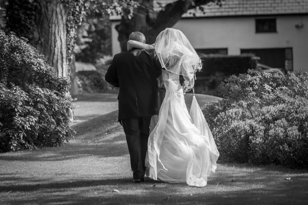 Black and white photo of a bride and groom walking arm in arm along a grassy path with bushes and trees around them.
