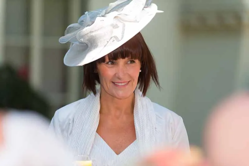 Woman smiling, wearing a white dress and a large white decorative hat with fabric details.