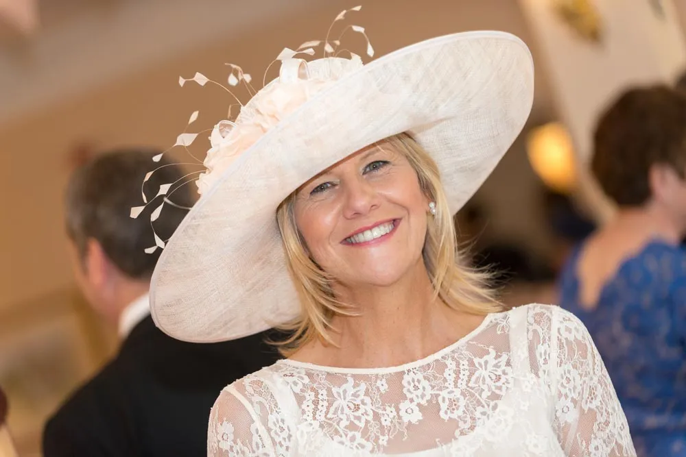 Smiling woman wearing a large white decorative hat and a white lace dress at an indoor event.