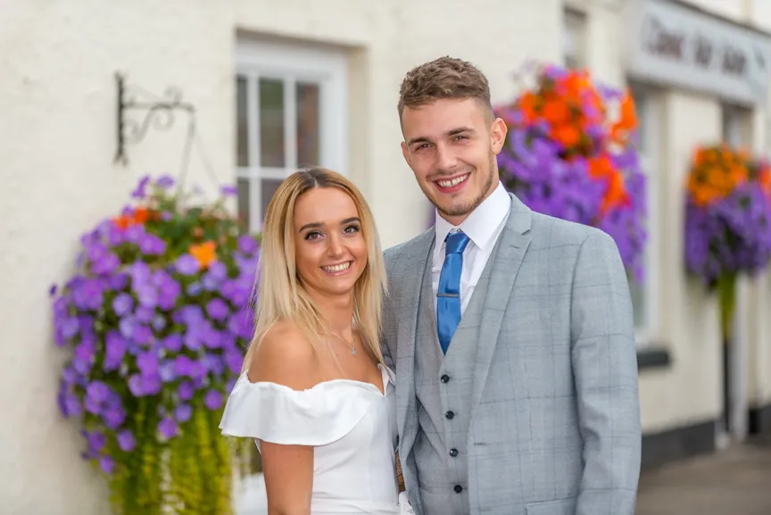 Smiling couple dressed in wedding attire posing outdoors with hanging flower baskets in the background.