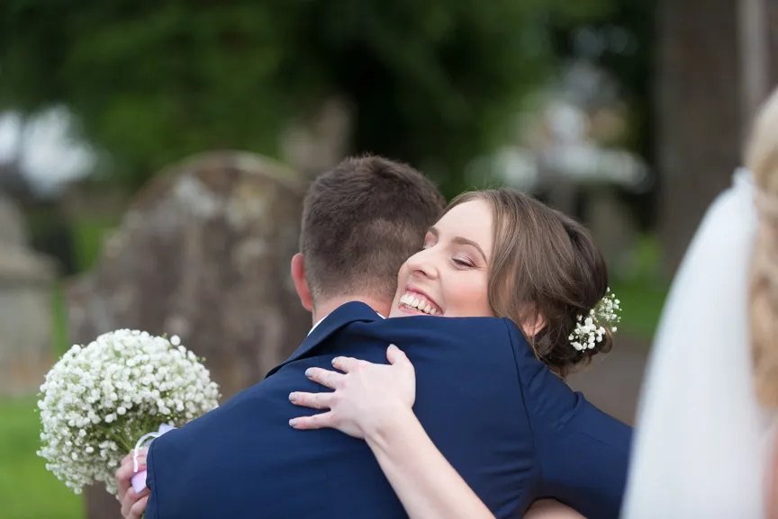 Bride with baby’s breath flowers in hair smiling and hugging groom in navy suit outdoors.